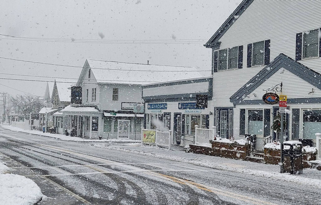Snow falling on a small town street with snow-covered road and storefronts at midday.”